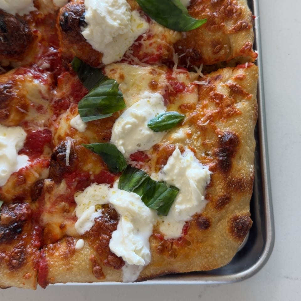 Close-up of square pizza topped with tomato sauce, melted cheese, creamy mozzarella, and fresh basil in a metal tray.